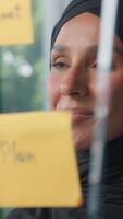 sorridente árabe muçulmano o negócio mulher olhando em adesivos em vidro dentro moderno escritório empresária dentro hijab profissional fêmea líder desenvolvedor lendo pegajoso notas sorridente projeto analisando gerenciando video