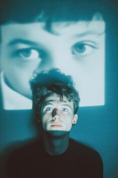 Young man gazes thoughtfully at home movie projected on wall, illuminated by soft light, creating a nostalgic atmosphere of reflection and memory photo