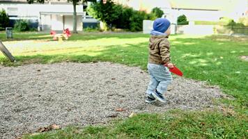 liten pojke löpning på de gräs på de lekplats. barn spelar i de höst på de lekplats. litet barn i en jacka och hatt i blåsigt väder på en promenad. härdning av de barnets kropp. video