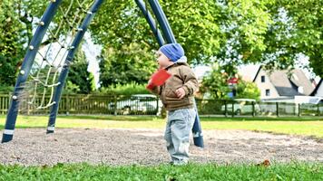 pojke löpning på de gräs på de lekplats. barn spelar i de höst på de lekplats. litet barn i en jacka och hatt i blåsigt väder på en promenad. härdning av de barnets kropp. video
