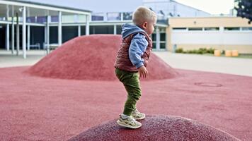 en 2-åring pojke i en väst och gymnastikskor kör runt om de lekplats och spelar under en promenad. litet barn åtnjuter en värma höst dag på de lekplats. barn utveckling. video