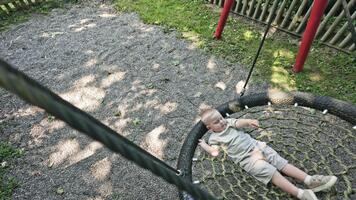 A 2 year old toddler in shorts swings on a spider on a warm summer day. A toddler boy swings on a playground in a place. Entertainment for a child on a playground video
