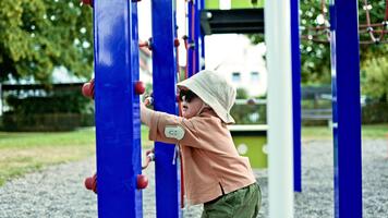 allegro bambino piccolo nel occhiali da sole giocando su terreno di gioco. bambino nel Panama cappello su testa in esecuzione su terreno di gioco nel città parco. bambino esplorando ambiente bambino giocando indipendentemente su terreno di gioco video