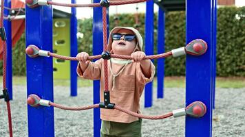 allegro bambino piccolo nel occhiali da sole giocando su terreno di gioco. bambino nel Panama cappello su testa in esecuzione su terreno di gioco nel città parco. bambino esplorando ambiente. bambino giocando indipendentemente su terreno di gioco. video