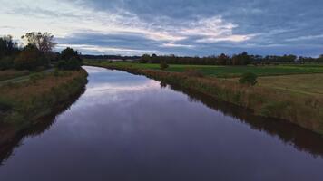 lugna flod flöden försiktigt genom fält under en skymning himmel fylld med moln och en ledtråd av solnedgång glöd video