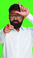 Indian man makes an L sign on his forehead with fingers while smiling playfully, teasing viewer as loser. Arabian guy isolated on green chroma key background showing joke, sarcasm and mock gesture. video