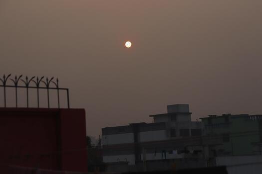 Sun setting over buildings with a hazy sky and a fence in the foreground photo