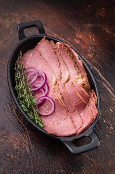 Thin sliced pastrami served in a cast iron pan with red onion rings and fresh thyme on a rustic dark textured background. Smoked deli beef arranged for cooking, serving or menu design. photo