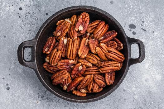 Brown pecan halves displayed in a cast iron pan resting on weathered wood against a grey textured background creating a natural moody food visual photo