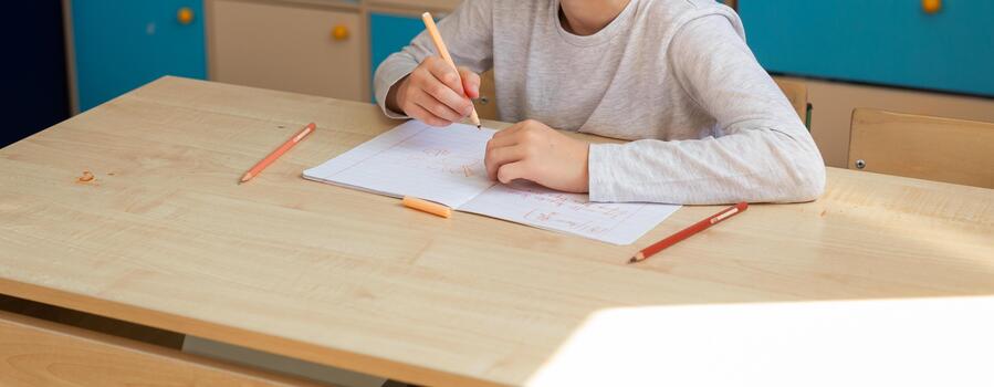 Child drawing in notebook with orange pencil during classroom lesson, banner with copy space. Creativity, imagination and focus on self-expression through learning. photo