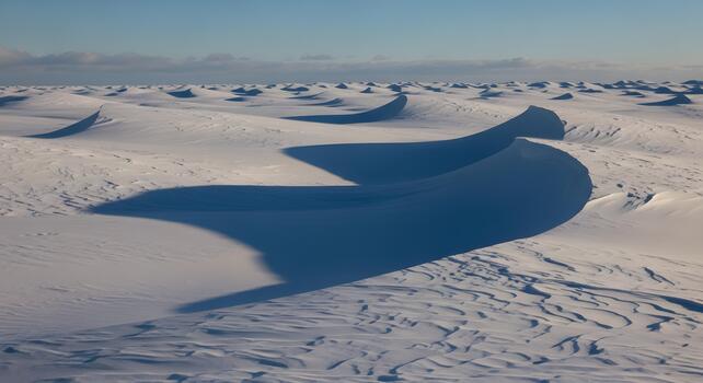 Abstract patterns of wind-sculpted snow dunes stretch across a pristine polar landscape under a clear winter sky, highlighted by dramatic long shadows creating a serene and vast cold environment photo