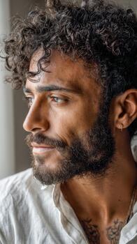 Young man with curly hair and beard looking thoughtfully out of a window photo