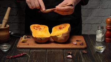 A chef skillfully halves a ripe squash and adds spices using a small spoon. The warm, rustic kitchen is filled with cooking ingredients and tools, creating an inviting atmosphere. video