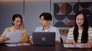 tre asiatisk individer förlova sig i en allvarlig diskussion på en tabell. varje person recensioner grafer och statistik visas på bärbara datorer, i samarbete analyserar data med fokus och avsikt. video