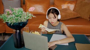 asiatisk kvinna studerande åtnjuter lyssnande till lugnande musik genom hörlurar medan uppslukad i läsning en lärobok och och använda sig av en bärbar dator dator till hitta information i henne inbjudande studie miljö video