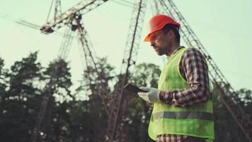 Engineer in hardhat is using tablet computer in power station. Inspecting worker using digital tablet with high voltage tower background. Electrician measures current level, power grid for supply video