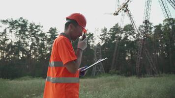 elektrisk ingenjör på hög Spänning kraft transformator substation rapportering problem med walkie talkie och utseende in i urklipp. professionell elektriker med bärbar radio nära hög Spänning torn video