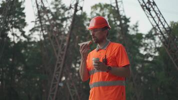 Engineer having lunch near power lines and electricity pylons in the field. An electrical substation worker in a protective helmet eats a sandwich and drinks coffee during a break at the workplace. video