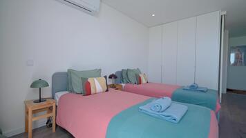 Sunlight illuminates a modern minimalist bedroom. Featuring two single beds adorned with pink and teal bedspreads. Complemented by stylish geometric patterned pillows and a bedside table with a lamp video