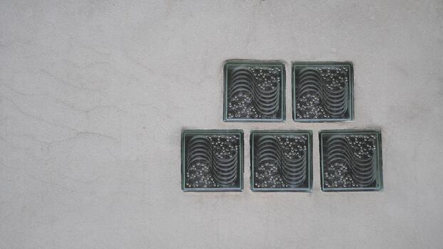 Five square, patterned glass blocks are embedded in a rough, gray concrete or stucco wall, forming a small decorative window or vent, with ample negative space on the left. photo