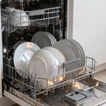 Open dishwasher with clean wet plates stacked in the lower rack and cups in the upper rack, water droplets visible on glossy porcelain. Proper loading and household cleaning routine photo