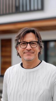 A man wearing glasses and a sweater standing in front of a house photo