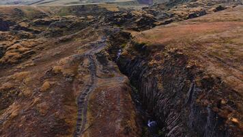 Aerial view follows a small stream through Sigoldugljufur in Iceland, showing basalt columns, small falls, a rim footpath with rope, and autumn tones under soft light. video