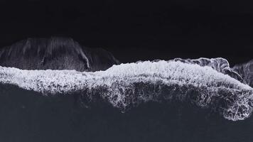 Top down aerial view shows black sand and white surf on an Icelandic shore near Vik. Waves roll and break as the camera drifts parallel, forming lace like foam patterns. video