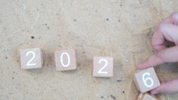 stänga upp av trä- block med tal 2025 vändning till 2026 på en sand bakgrund, betona de begrepp av tid progression och de övergång av år. video