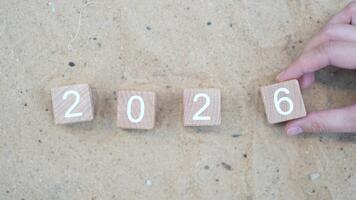 Hand placing wooden blocks with numbers 2026 on sandy beach background, symbolizing a transition into the new year and fresh beginnings ahead. video
