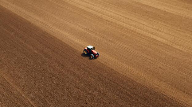 An aerial view of a blue and red tractor plowing an expansive brown field under bright sunlight. photo