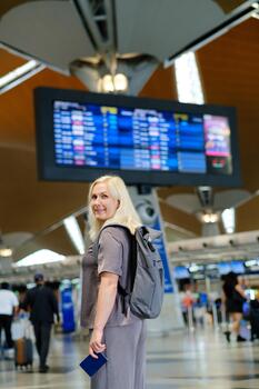 Woman waiting at the airport terminal with a backpack, smiling at the camera while flight information displays are visible in the background. photo