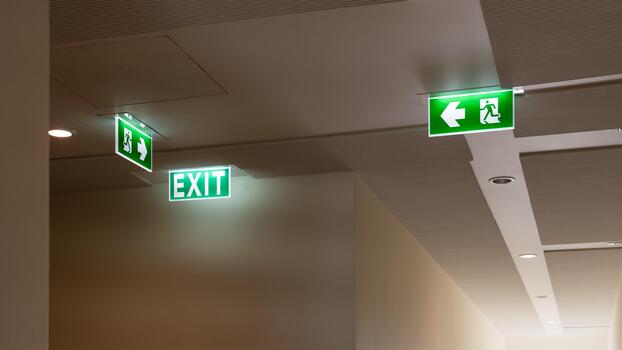 A green fire escape sign hangs from the ceiling, showing an exit sign green light box with a running man and an arrow pointing right for safety and evacuation.a photo