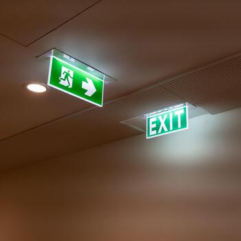 A green fire escape sign hangs from the ceiling, showing an exit sign green light box with a running man and an arrow pointing right for safety and evacuation.f photo