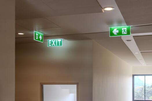 A green fire escape sign hangs from the ceiling, showing an exit sign green light box with a running man and an arrow pointing right for safety and evacuation.c photo