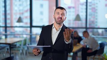 allvarlig man i kostym samtal i främre av kamera som visar grafer. självsäker manlig högtalare utför presentation i kontor. människor arbete på de skrivbord på bakgrund i fläck. video
