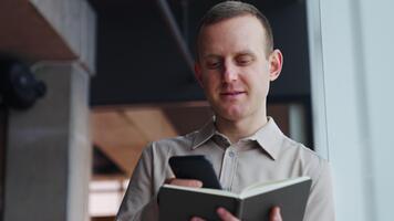 maturo uomo detiene il taccuino ha aperto nel davanti di lui e Stampa qualcosa su il Telefono. lui lanci interessato Guarda in il finestra. video