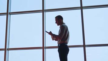 hombre sentado a el escritorio, mirando a su teléfono, hablando y sonriente a alguien en el habitación. bajo ángulo vista. panorámico ventanas a fondo video