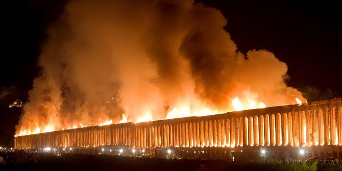 A massive building with columns burns brightly with huge flames and smoke during a nighttime event watched by a crowd photo