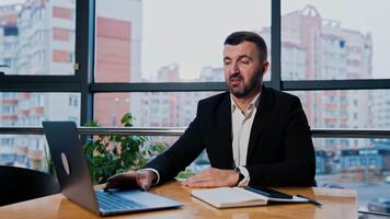 affärsman använder sig av bärbar dator Sammanträde på de skrivbord nära de stor fönster. man trycker på nycklar på dator och samtal till kamera. stadsbild på bakgrund. video