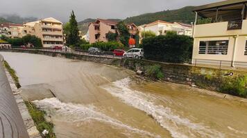hinchado lodoso corriente corriendo pasado orilla edificios, blanco espuma cascada terminado pequeño gota, nublado colinas en fondo, turbulento fluir que lleva limo video