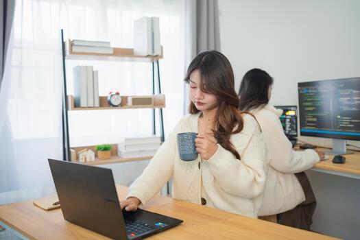 Young women working in a modern office environment, one enjoying coffee while using a laptop, the other focused on coding at a computer station photo
