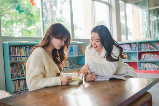 Two young women engaging in conversation while studying in a cozy library setting, surrounded by books and natural light, sharing learning experiences. photo