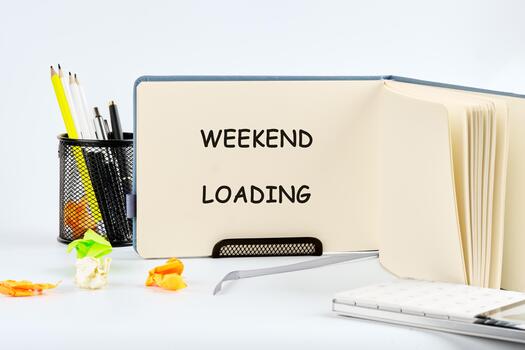 An open notebook displays the phrase WEEKEND LOADING. Nearby are writing implements, a laptop, and discarded, crumpled papers, all set against a plain, bright background photo