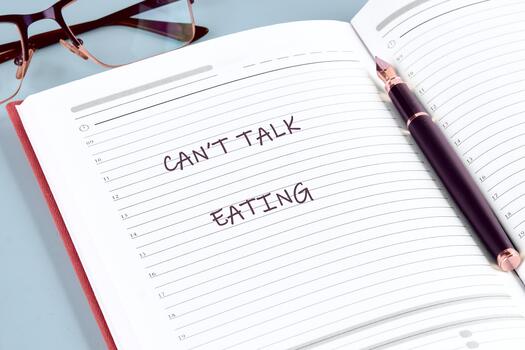 Open notebook with the words Cant Talk Eating hand-written on a lined page. A pen rests on the book with glasses in the background on the table photo