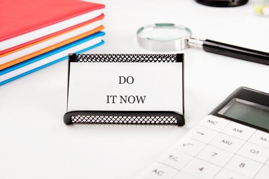 A card reading DO IT NOW sits on a desk with stacked colorful books, a magnifying glass, and a calculator, promoting instant task completion photo