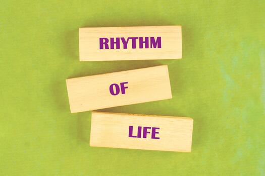 Three light-colored wooden blocks are stacked. Each block has a word in purple letters forming the phrase RHYTHM OF LIFE on a bright, textured, green background photo