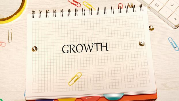 A grid notebook lies open on a desk, boldly displaying the word growth. Colorful paper clips and a magnifying glass scattered around add a touch of creativity to the space photo