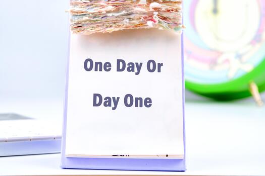 A motivational calendar sits on a clean desk, displaying the phrase One Day Or Day One. The colorful clock in the background adds a cheerful touch, encouraging action and reflection photo