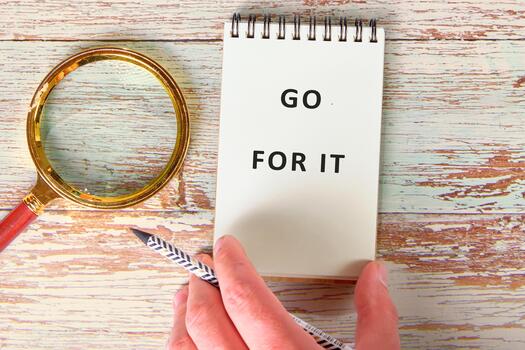 A hand grips a patterned pencil ready to write in a simple notebook that features a motivational phrase urging to take action. A shining magnifying glass rests nearby on the tables surface photo